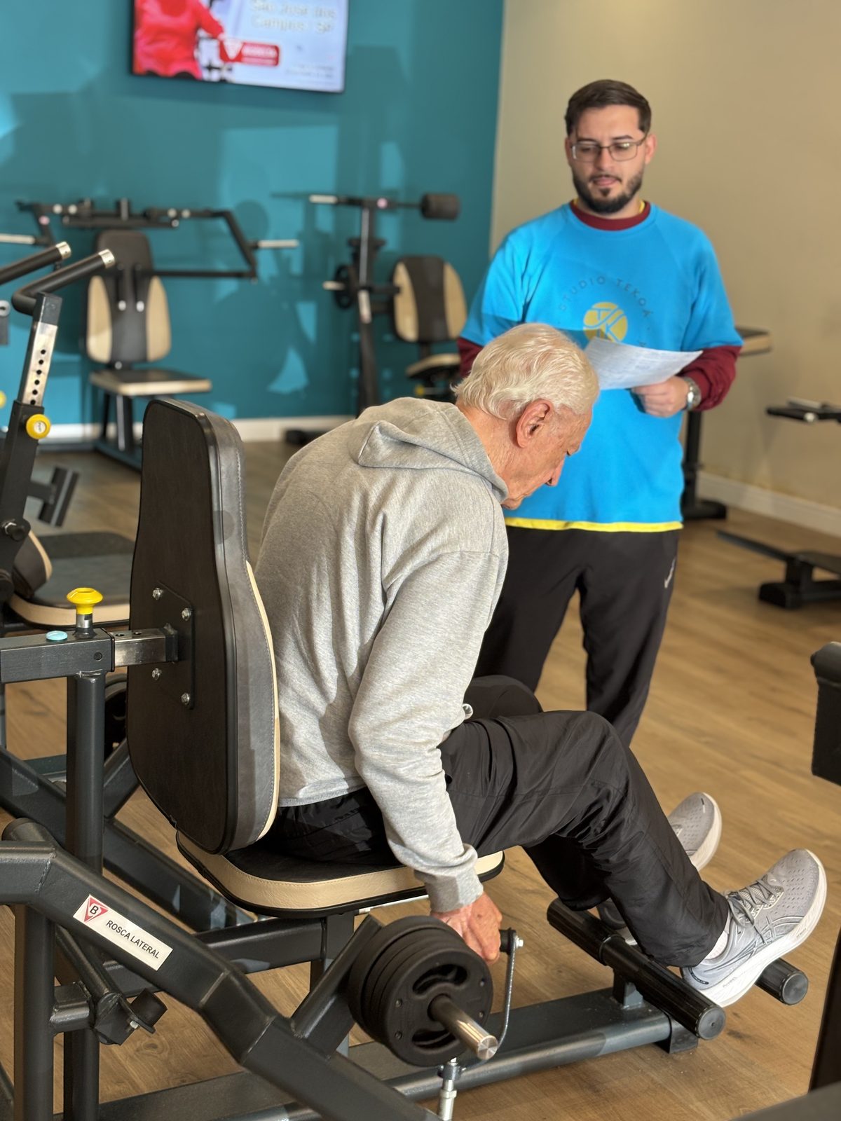 A member training on a weight machine while a Studio Tekoá professional reviews his training sheet