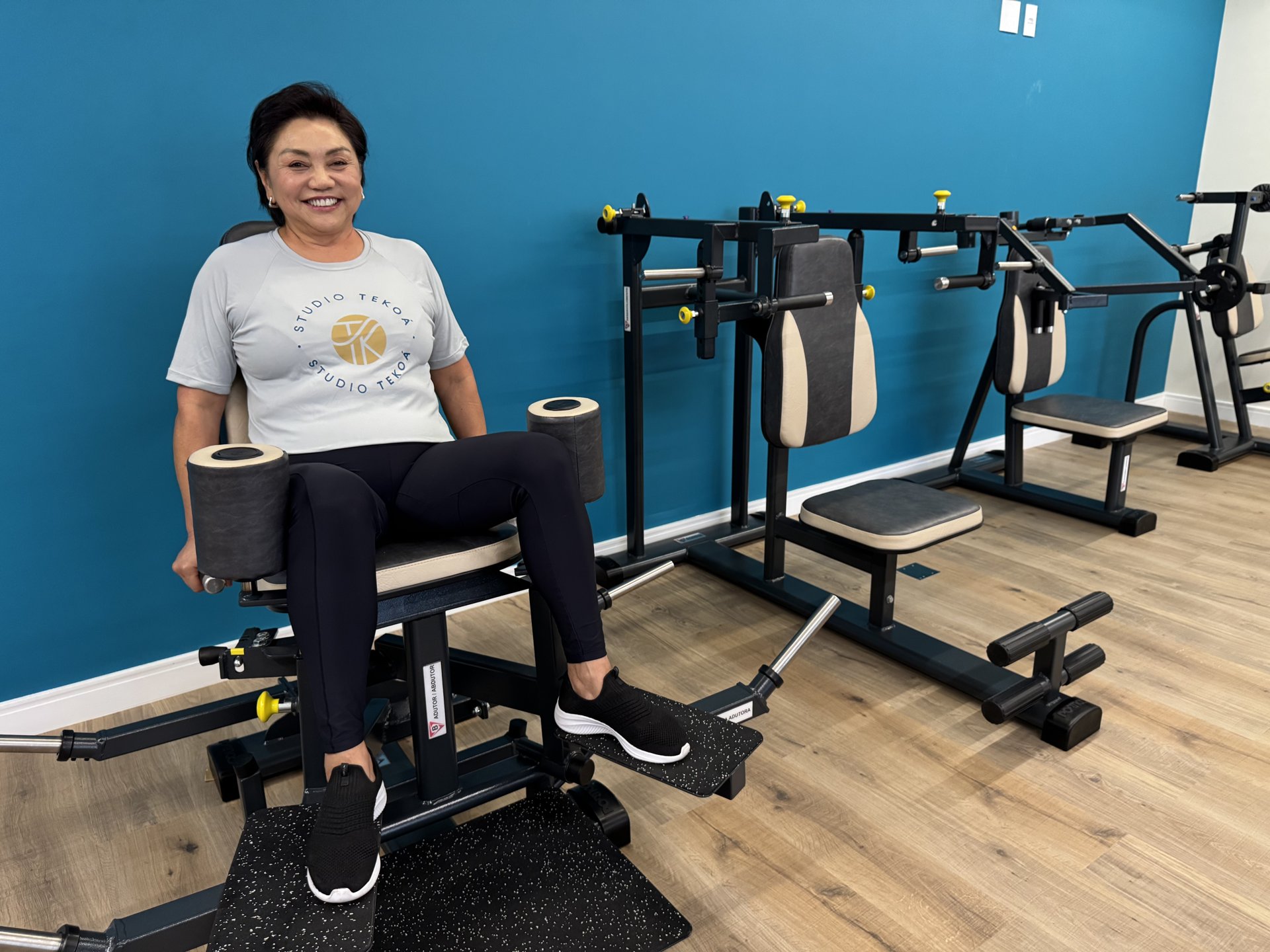 A smiling woman in a Studio Tekoá t-shirt sits on a strength machine in the studio, with a blue wall and training equipment behind her