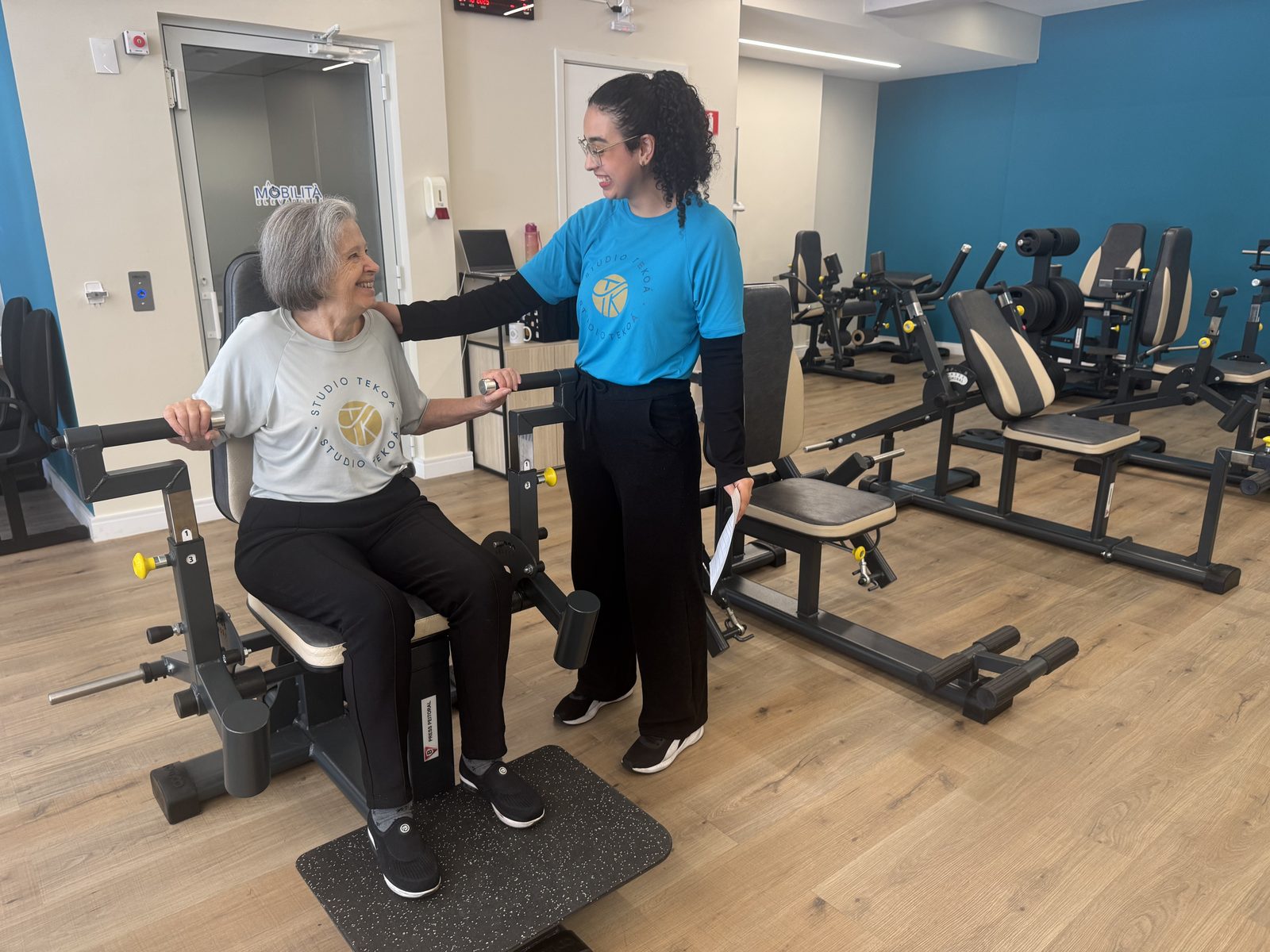 A smiling member on a chest press machine while a Studio Tekoá professional stands beside her with a hand on her shoulder